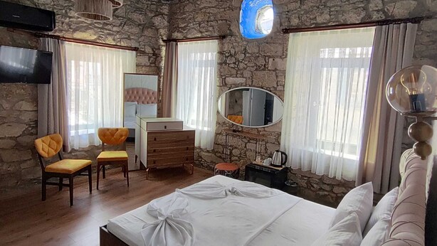 A bedroom with a stone wall, a bed with white linens, a wooden dresser, a chair, a mirror, and a hanging light fixture. - Baba Evi Alaçatı (Cesme)