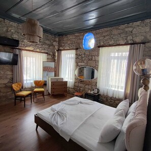 Quarto com parede de pedra, cama com roupa de cama branca, cómoda de madeira, cadeira, espelho e luminária pendente.