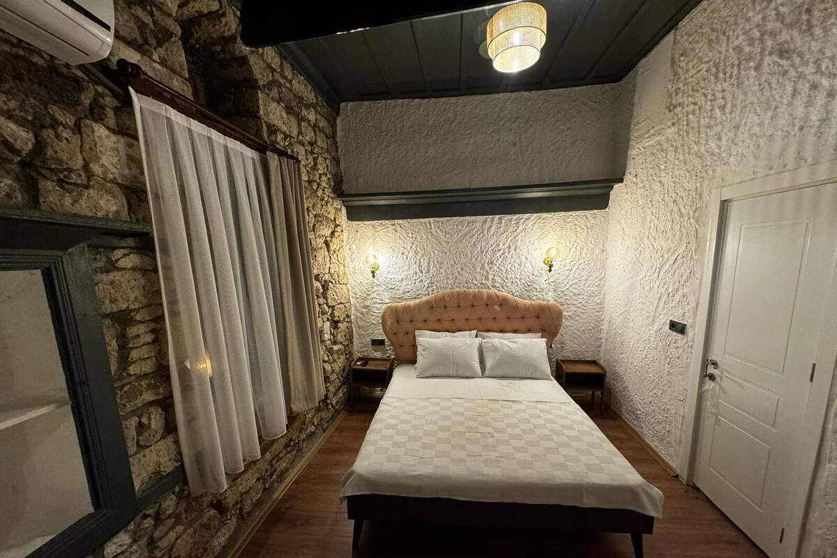 A bedroom with a stone wall, a checkered bedspread, a tufted headboard, two bedside lamps, and a door.