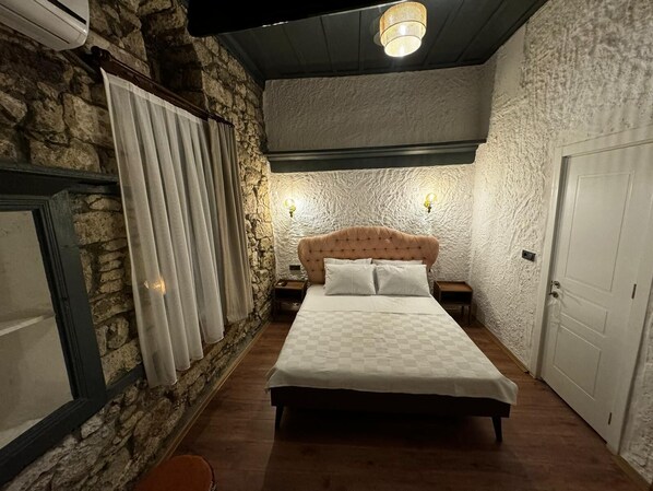 A bedroom with a stone wall, a checkered bedspread, a tufted headboard, two bedside lamps, and a door. - Baba Evi Alaçatı (Cesme)