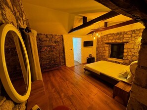 A bedroom with a stone wall, wooden floor, a bed, a TV, and a fireplace. - Baba Evi Alaçatı (Cesme)