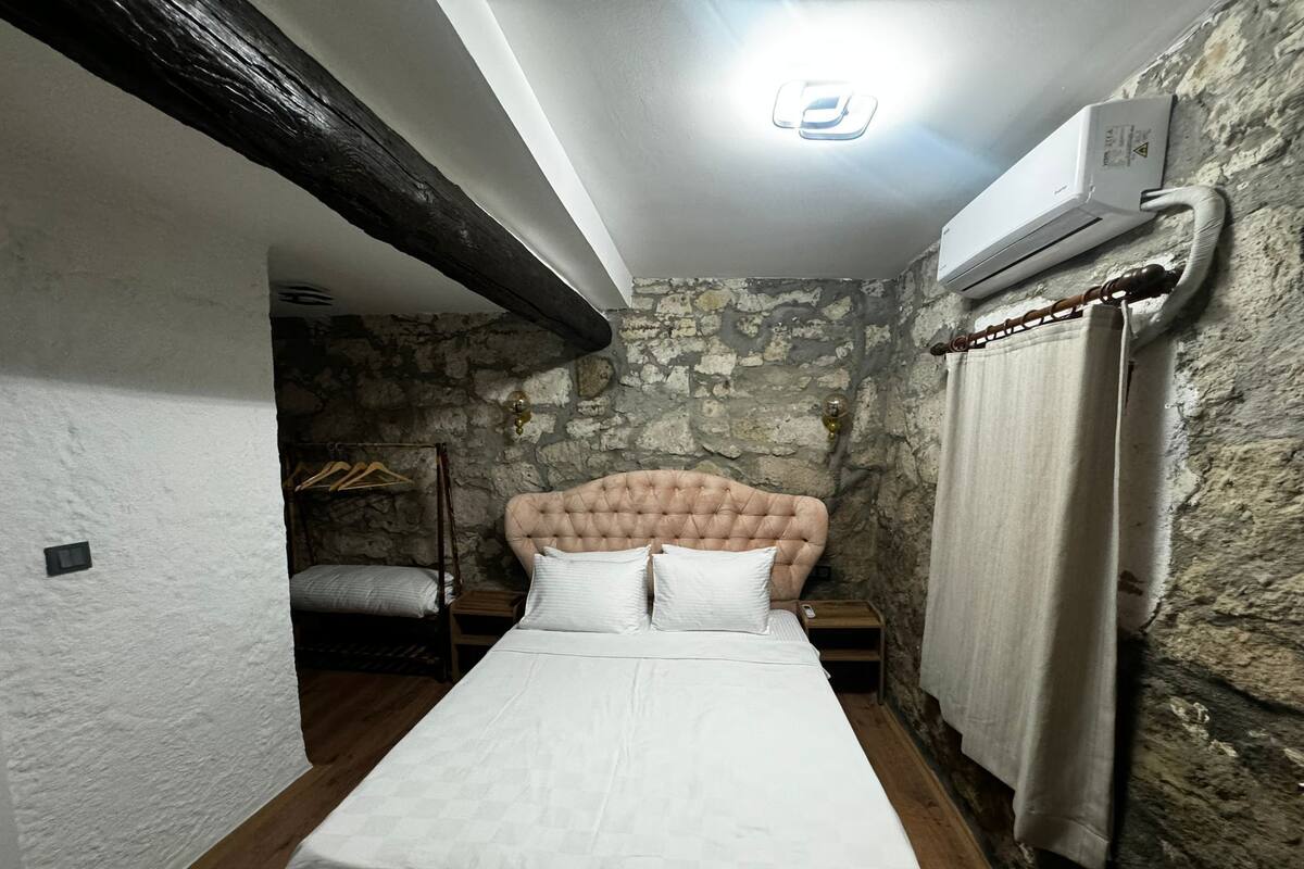 A narrow room with a bed, a wooden bench, a wall-mounted heater, and a stone wall.