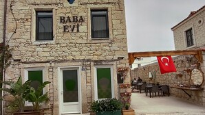 A stone building with a sign reading "BABA EVI". There are two visible windows, a door, and a small balcony. The building has a rustic appearance with a tiled roof. - Baba Evi Alaçatı (Cesme)