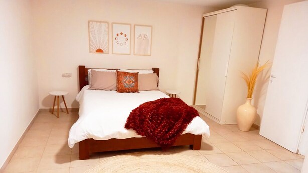 A bedroom with a bed, a round rug, a small table, and a vase with dried plants. - Spacious, Peaceful 2BD, Mamad, Garden, Parking near Kfar Sava (Gan Haim)