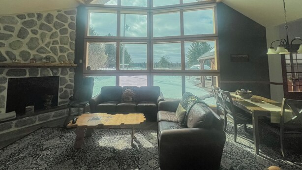 A cozy living room with a stone fireplace, a large window offering a view of the outdoors, a leather sofa, and a dining area with a table and chairs. - Peaceful Getaway on Mio Pond (Mio)