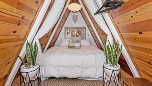 Una habitación de madera acogedora y triangular con una cama, dos plantas en macetas y una lámpara colgante.