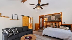 Un dormitorio moderno con cama, sofá y una mesita. Se ve un ventilador de techo, una puerta de madera y una cocina a través de un umbral abierto.