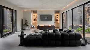 Un soggiorno moderno con un ampio divano modulare, una poltrona, un tavolino da caffè e una TV a schermo piatto montata a parete. Si notano mensole con oggetti decorativi e una pianta in vaso.