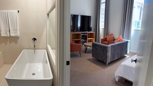 Une chambre d’hôtel moderne avec une grande baignoire blanche, un téléviseur à écran plat, un canapé gris et un lit avec une literie blanche.