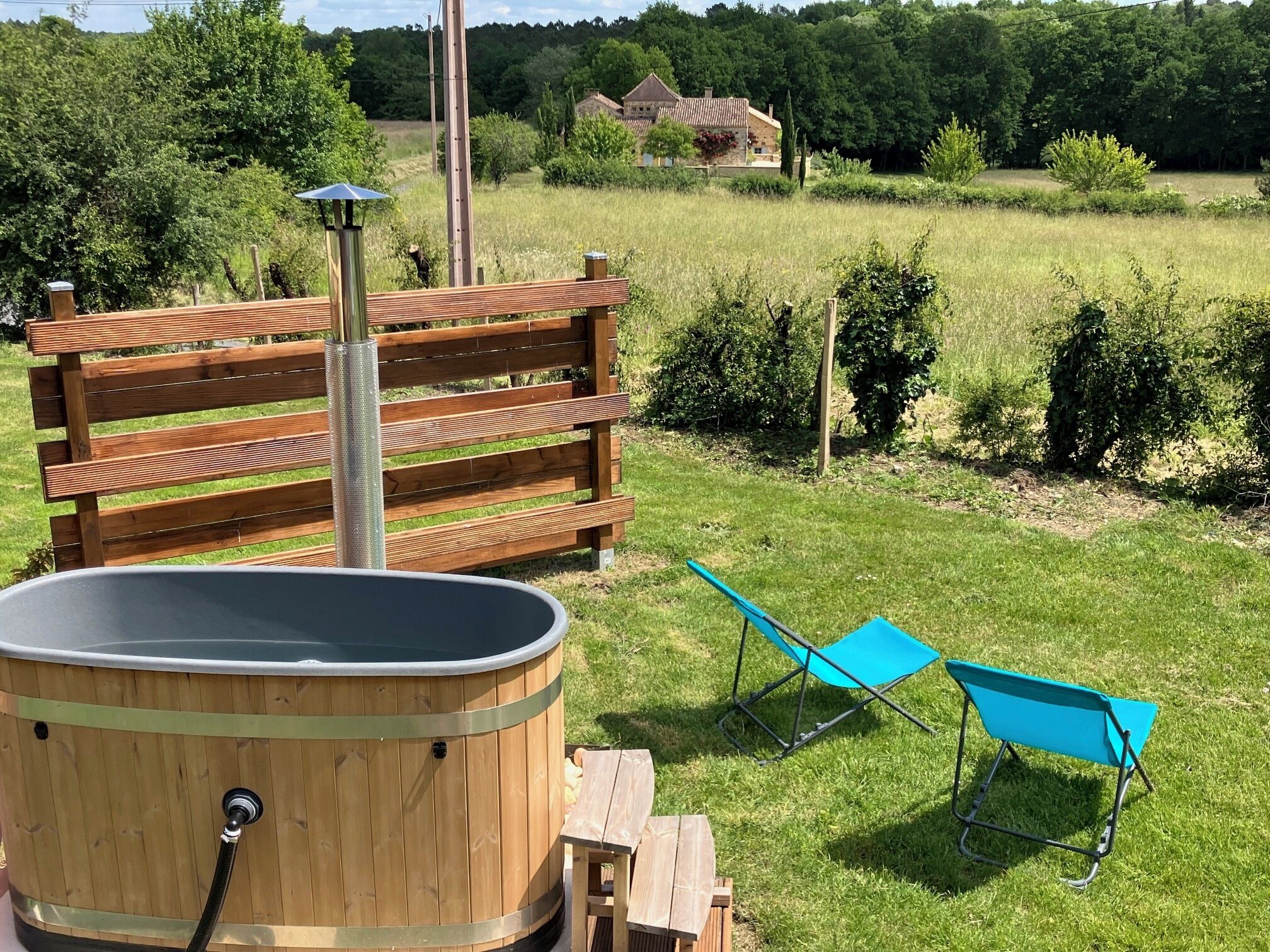 Ein hölzernes Whirlpool mit Metallstange und blauem Klappstuhl in einer grünen Außenanlage.
