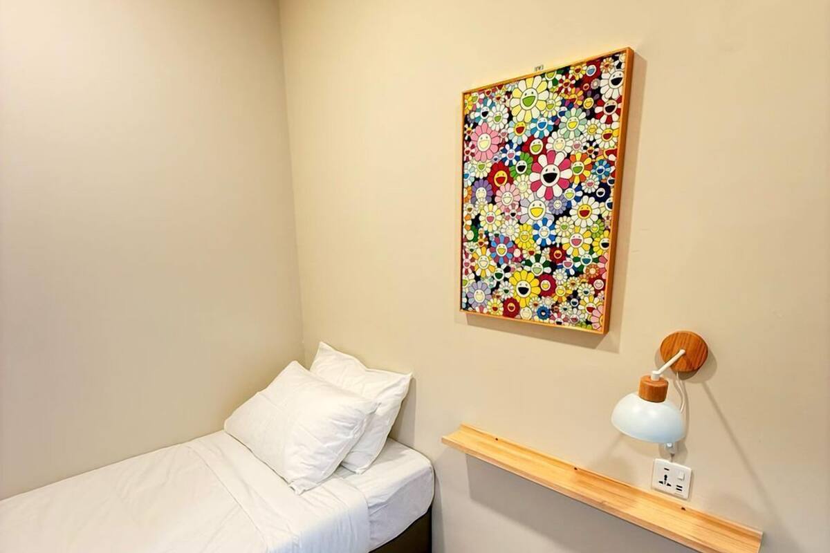 A small, neatly arranged bedroom with a single bed, a wooden floor, a wall-mounted shelf with a lamp, and a colorful floral artwork.