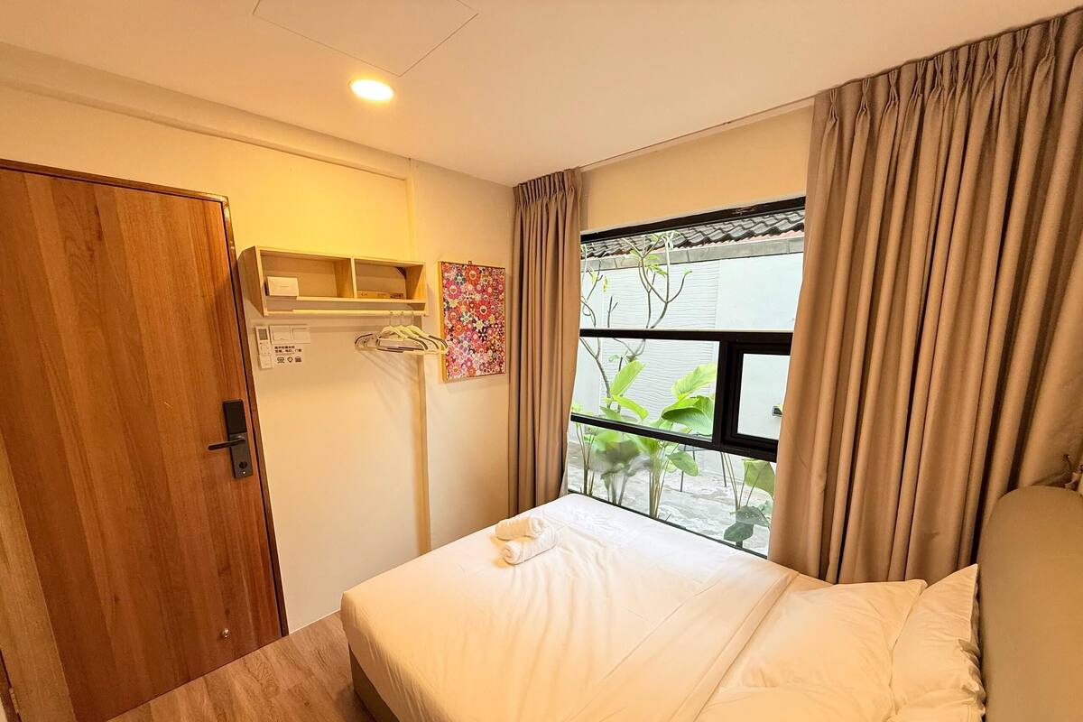 A hotel room with a bed, a wooden door, a window with curtains, and a small shelf with a floral decoration.