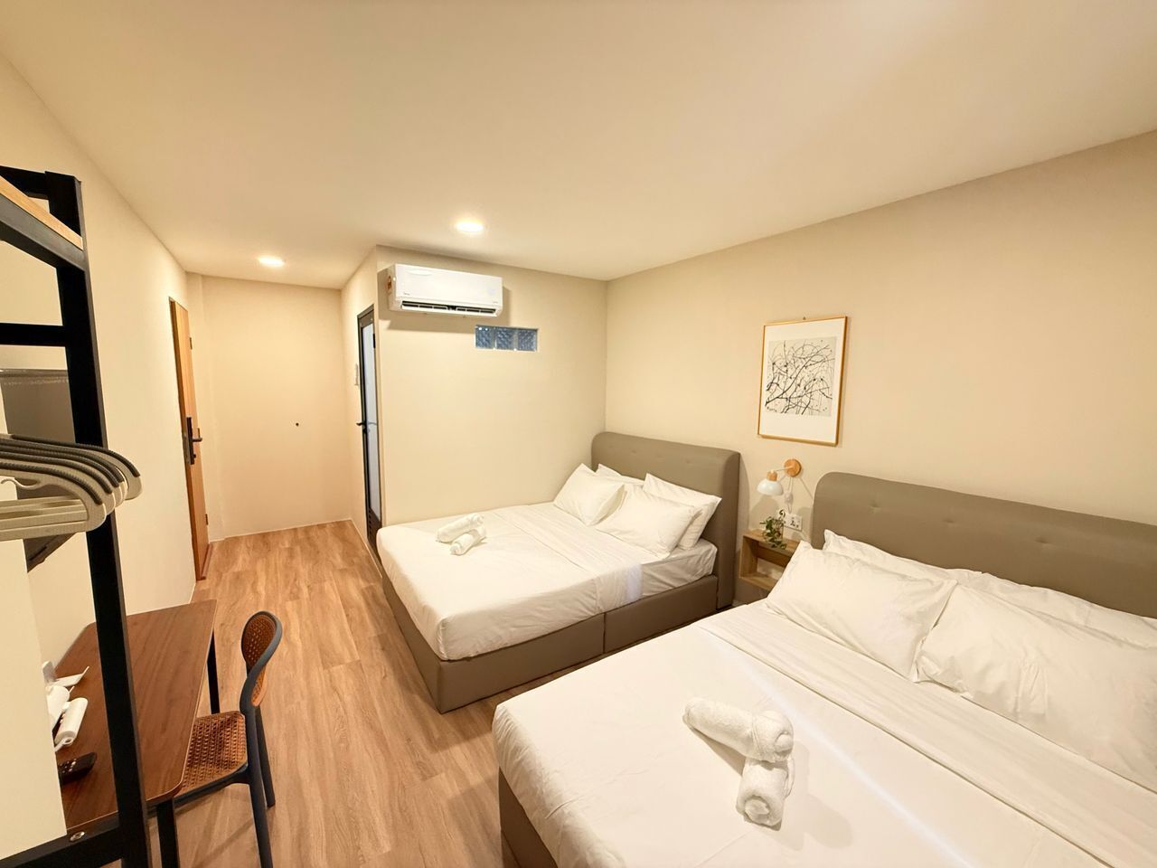 A hotel room with two beds, a wooden floor, a desk with a chair, an air conditioning unit, and a framed picture on the wall.