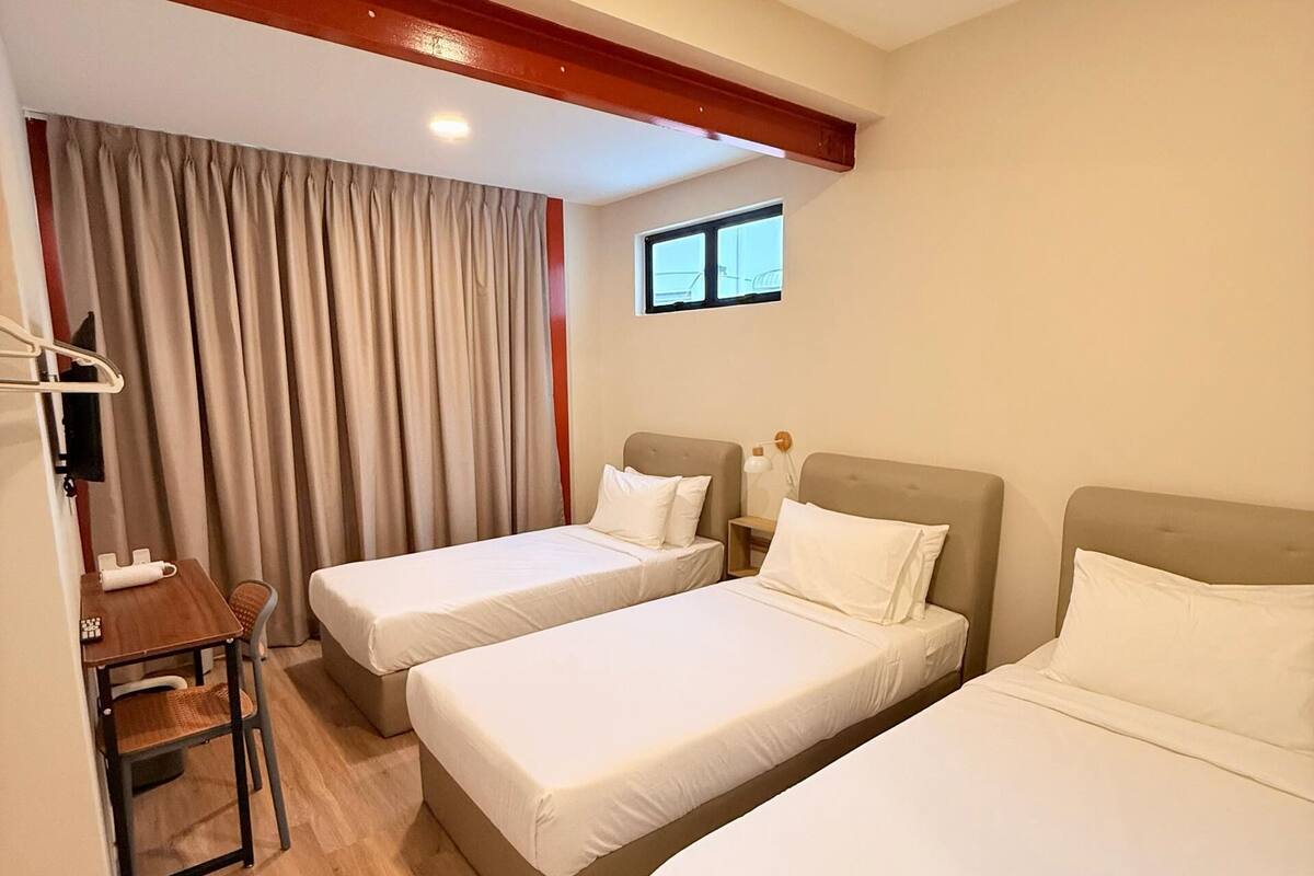 A hotel room with two single beds, a small table with a chair, and a window with curtains.