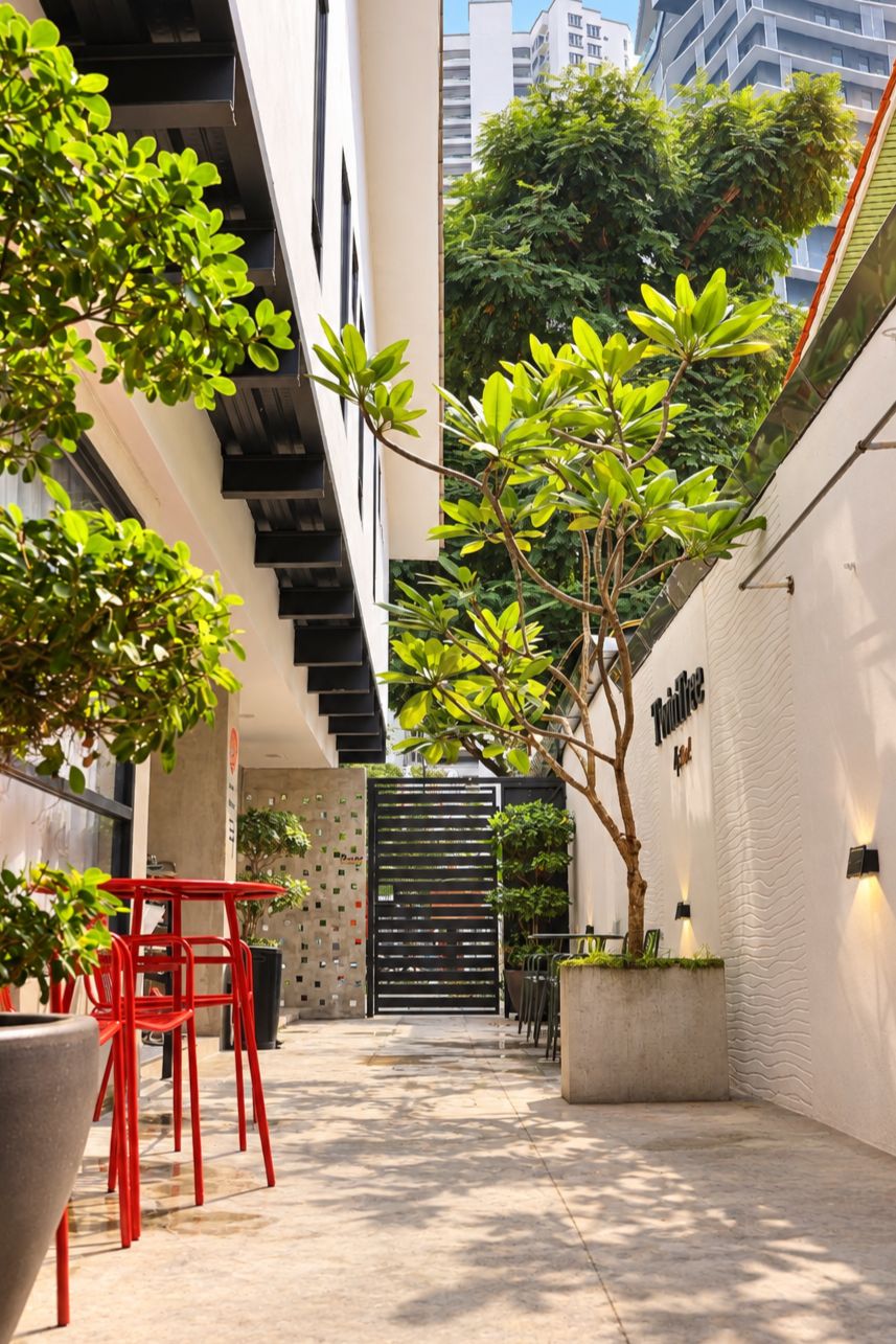 Un corridoio esterno stretto con un edificio moderno su un lato, un vialetto alberato e una zona relax con sedie rosse.