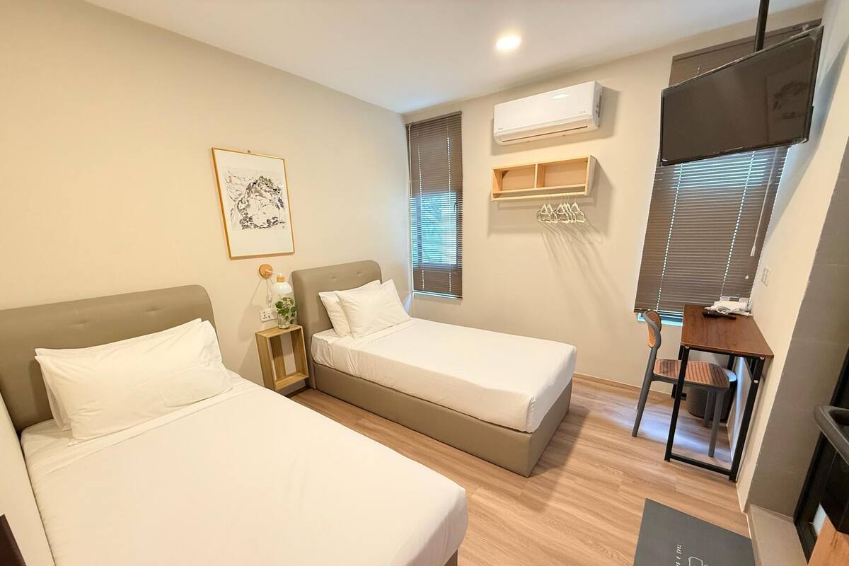 A hotel room with two beds, a desk, a chair, and a small table. There is a window with blinds, an air conditioning unit, and a framed picture on the wall.