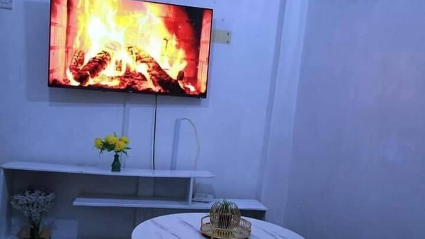 A living room with a TV mounted on the wall displaying a fire, a small round table with a remote, and a vase with flowers on a shelf below. - Lux Suites Ruby Court Apartments Kisumu (Kisumu)