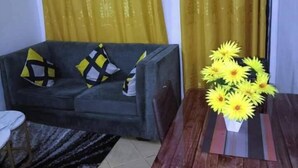 A living room with a grey sofa, yellow and black cushions, a wooden dining table with a vase of yellow flowers, and yellow curtains. - Lux Suites Ruby Court Apartments Kisumu (Kisumu)