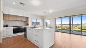 A modern kitchen with white cabinetry, a central island, and a large sliding glass door leading to a patio with a view of a residential area. - 29 Blanche Pd, Hindmarsh Island (Hindmarsh Island)