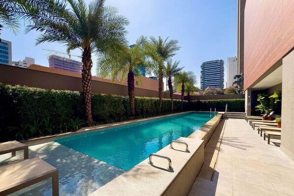 A swimming pool with palm trees, a clear sky, and modern buildings in the background.