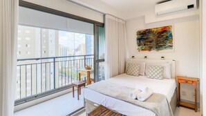 A modern hotel room with a large balcony, a bed with white linens, a small wooden table, and a colorful abstract painting on the wall. - Vitralli  (Sao Paulo)