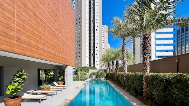 A swimming pool with lounge chairs and potted plants, surrounded by tall buildings and palm trees. - Vitralli  (Sao Paulo)