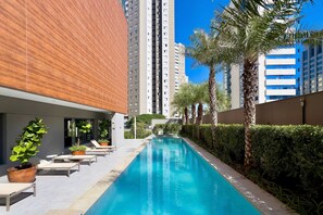 A swimming pool with lounge chairs and potted plants, surrounded by tall buildings and palm trees. - Vitralli  (Sao Paulo)