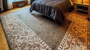 Een knus slaapkamer met een houten plafond, een groot bed met een donkere spreidekbed, twee bedlampjes en een raam met gordijnen.