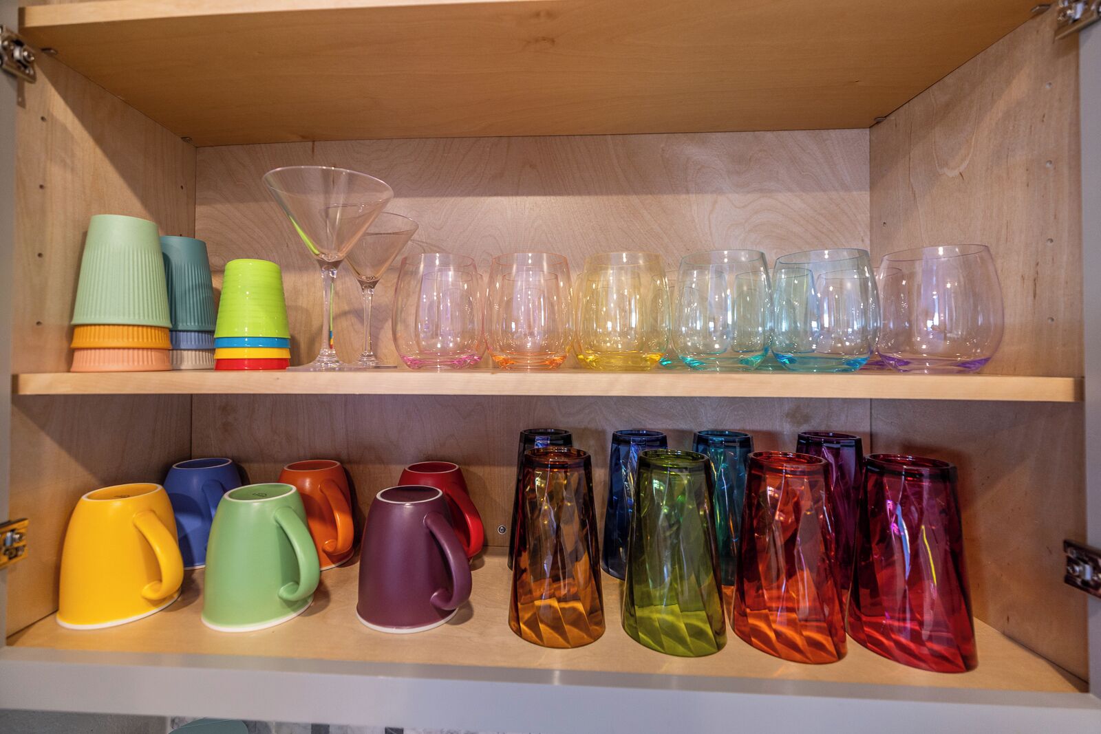 A wooden cabinet with shelves holding various colored glassware and mugs.
