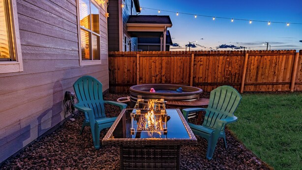 An outdoor patio with a fire pit table, two Adirondack chairs, a hot tub, and a wooden fence. - Near Airport: Game Room, Mini Golf, Cowboy Pool, Smart TVs & Air Purifiers (Commerce City)