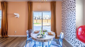 A dining area with a round table, clear chairs, and a view of a residential area through a large window. - Near Airport: Game Room, Mini Golf, Cowboy Pool, Smart TVs & Air Purifiers (Commerce City)