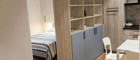 A modern, compact room with a bed, a built-in wardrobe with multiple shelves, a dining area with a table and chairs, and a kitchenette.
