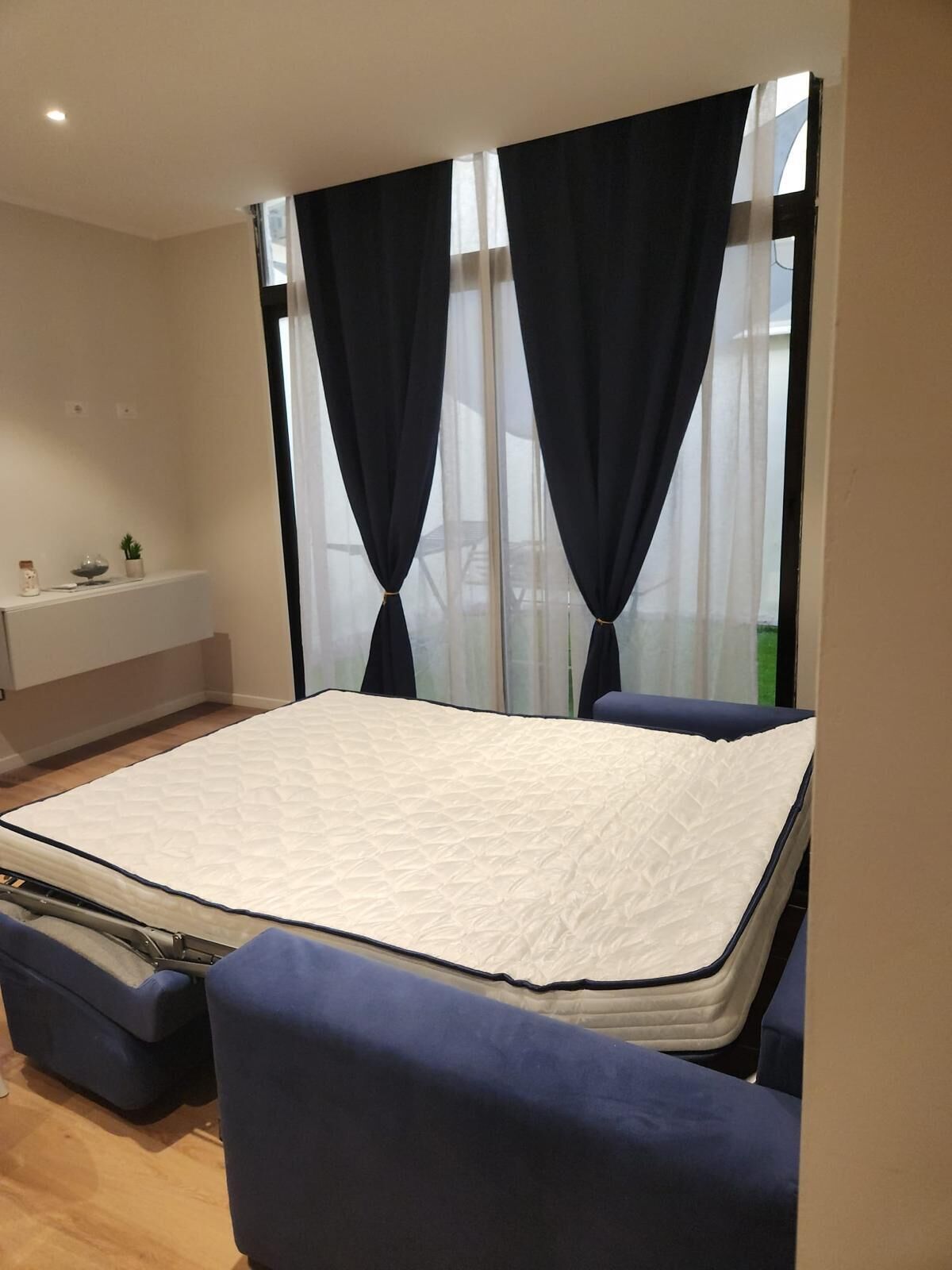 Una habitación compacta con cama, un pequeño rincón de estar, una ventana con cortinas y un estante en la pared con una planta.