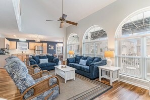 A spacious living room with a large window, a ceiling fan, and a kitchen in the background.