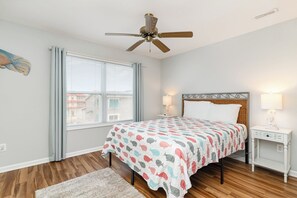 A bedroom with a bed, a nightstand, a ceiling fan, a window with curtains, and a view of buildings outside. - Don't Worry Beach Happy | Your Beach Getaway! (Ocean Isle Beach)