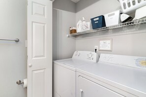 Une buanderie équipée d’un lave-linge et d’un sèche-linge à chargement frontal, d’une étagère métallique au-dessus pour ranger divers produits de lessive, et d’une porte dotée d’un porte-serviettes.