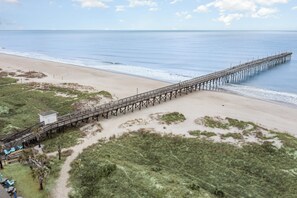 A beach with a wooden pier extending into the ocean, a small white building, and a grassy area with chairs. - Don't Worry Beach Happy | Your Beach Getaway! (Ocean Isle Beach)
