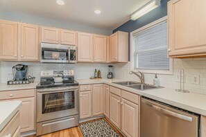 A modern kitchen with light wood cabinets, stainless steel appliances, a built-in microwave, and a window with blinds. - Don't Worry Beach Happy | Your Beach Getaway! (Ocean Isle Beach)