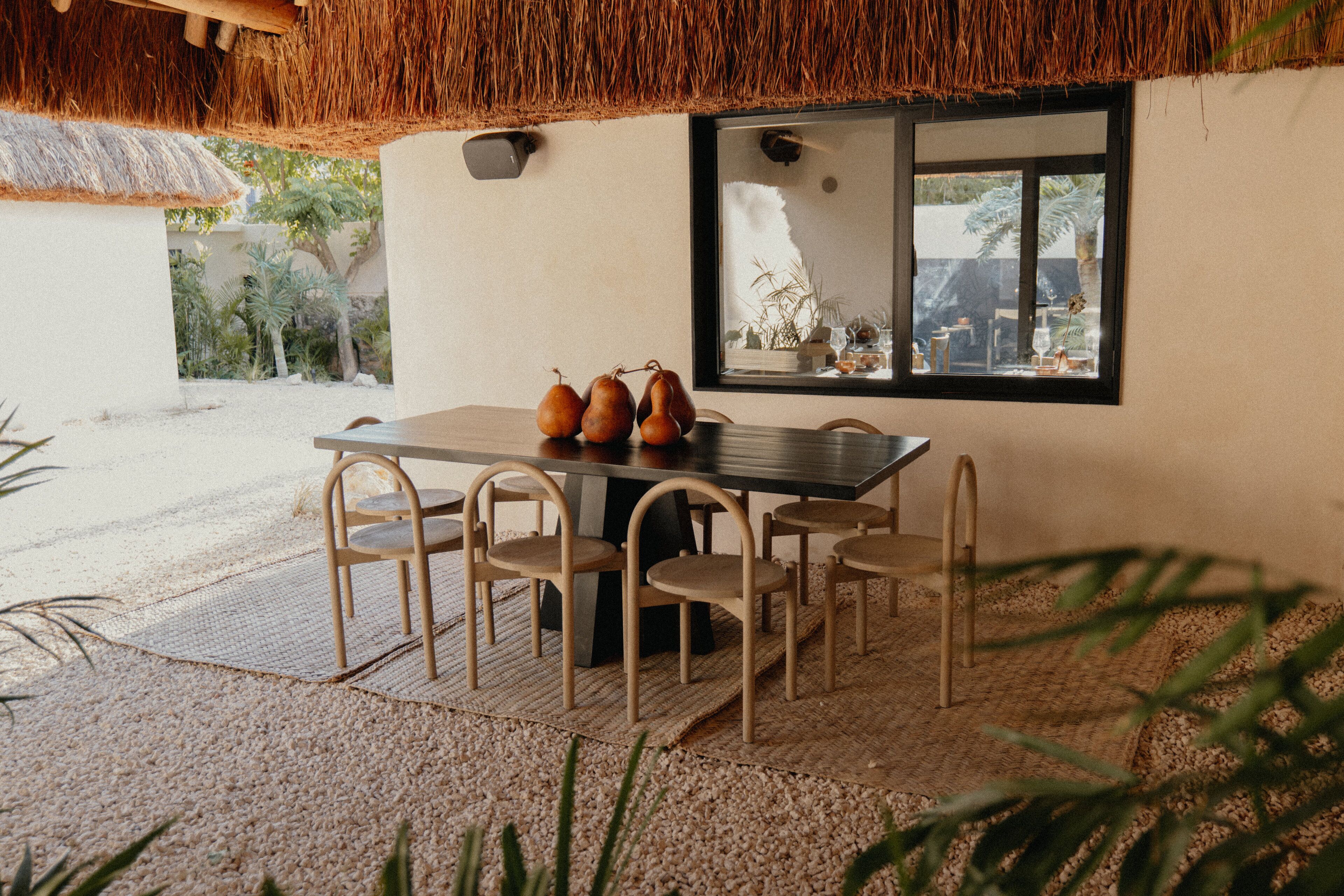 A dining area with a table and chairs, a thatched roof, and a window with a view of greenery.
