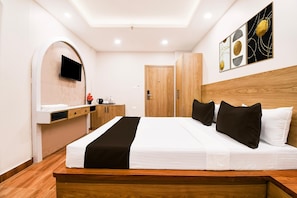 A modern hotel room with a wooden bed, a flat-screen TV mounted on an arched wall, a wooden wardrobe, and a small desk with a chair.