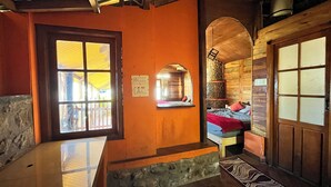 A cozy room with a wooden interior, a stone base, a bed with red bedding, and a window with a view. - Mount Olympus Hood cabin (Kodaikanal)