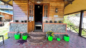 A wooden cabin with a stone wall entrance, a bed inside, and potted plants on the porch. - Mount Olympus Hood cabin (Kodaikanal)