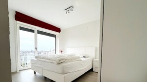 Une chambre moderne avec un grand lit, une table de chevet et une vue sur le paysage urbain, visible à travers une grande fenêtre.