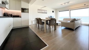 Une cuisine moderne avec des meubles blancs, un coin repas avec une table et des chaises, et un salon avec un canapé et de grandes fenêtres donnant sur l’océan.