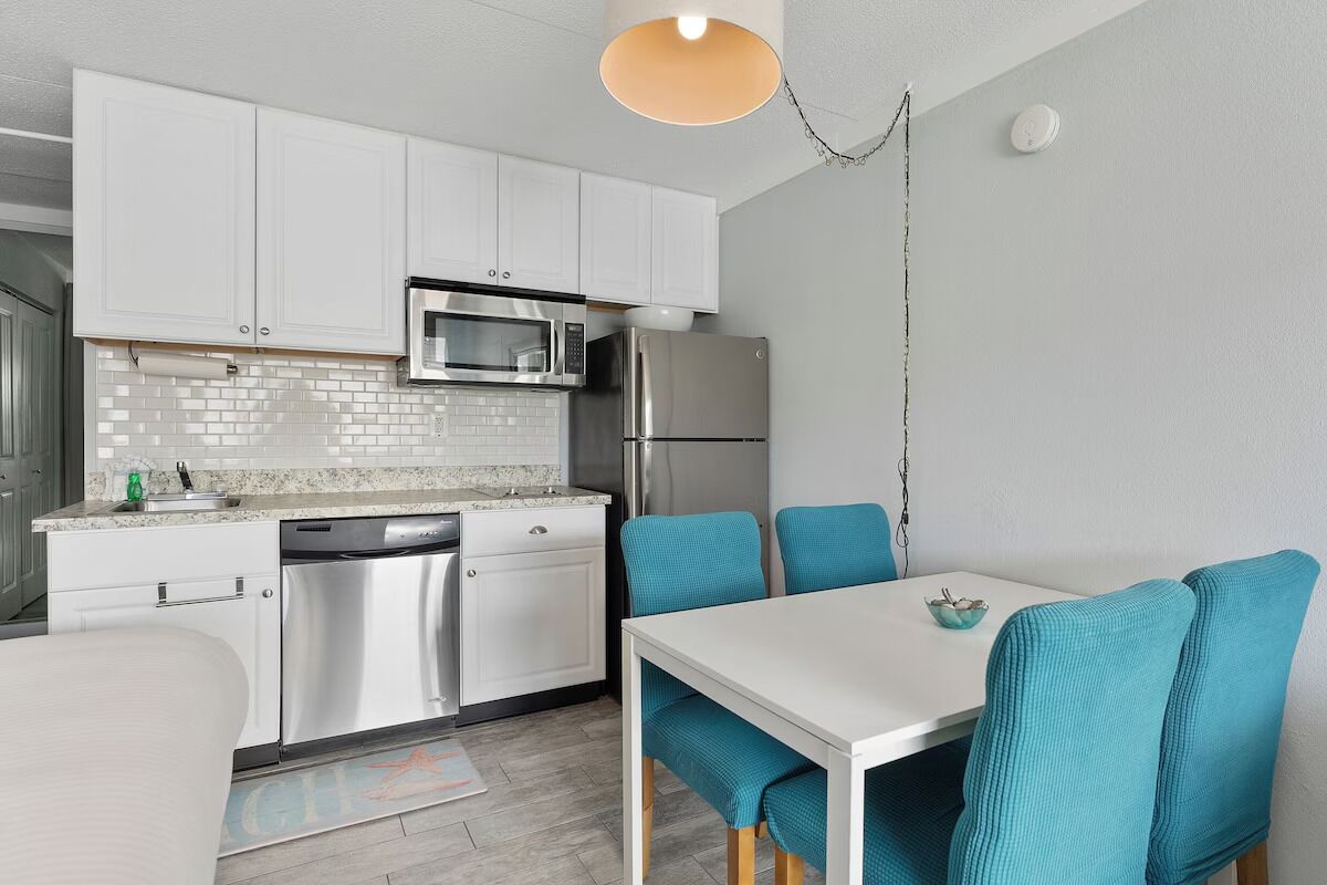 Une cuisine moderne avec des meubles blancs, un four à micro-ondes, un réfrigérateur et un coin repas agrémenté de chaises turquoise et d’une table blanche.