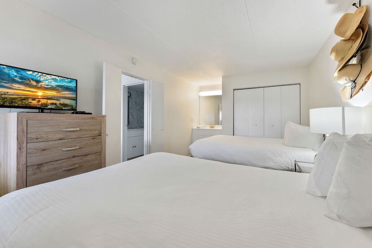 Ein Hotelzimmer mit einem großen Bett, einer Holzkommode, einem Fernseher, der einen Sonnenuntergang zeigt, und einem an der Wand befestigten Hutfänger.