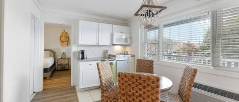 Cocina compacta con armarios blancos, microondas y comedor con sillas de mimbre.