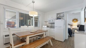 Cocina compacta con muebles blancos, microondas y refrigerador. Cuenta con una mesa de comedor con bancos y una ventana con persianas.