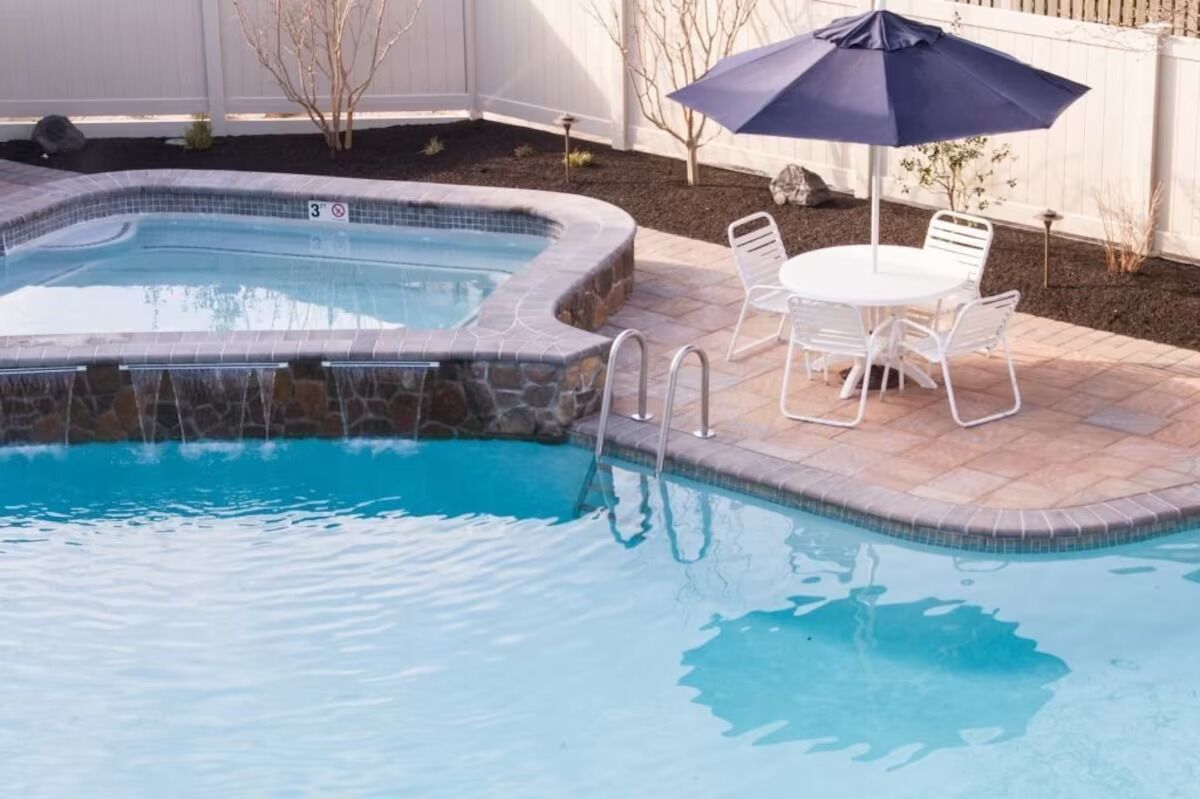 A swimming pool with a stone edge, a small waterfall feature, and a patio area with a table and chairs.