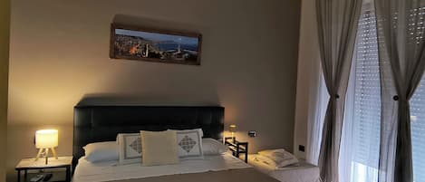 A hotel room with a large bed, two bedside tables, a nightstand, a painting on the wall, and a window with curtains.